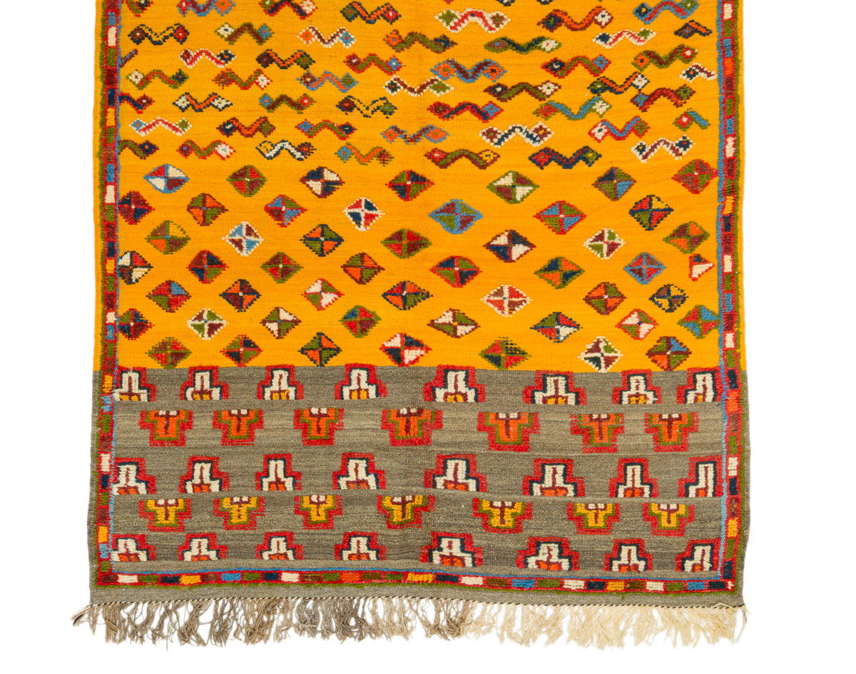 A kilim rug featuring a bright yellow-orange field with rows of multicolored diamond and zigzag motifs, a wide gray border with red, orange, and white geometric patterns, and cream-colored fringe along the bottom edge.