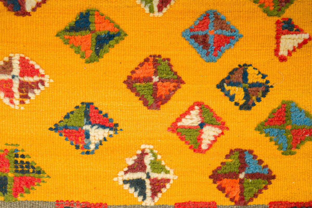 A close-up of a handwoven rug with a bright yellow background and repeating multicolored diamond motifs in red, blue, green, orange, and white.