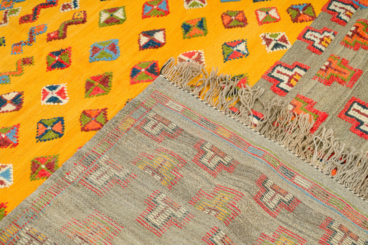 A close-up of a woven rug with two distinct patterned sections: a bright yellow section with multicolored geometric diamond motifs, and a larger gray section with red, beige, and white geometric patterns and fringed edges.