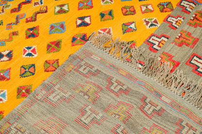 A close-up of a woven rug with two distinct patterned sections: a bright yellow section with multicolored geometric diamond motifs, and a larger gray section with red, beige, and white geometric patterns and fringed edges.