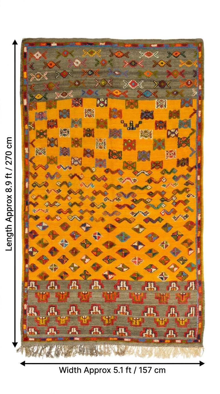 A flat-lay view of a rectangular woven rug with fringe, showing a mustard yellow center field with diamond and checker-like geometric motifs, bordered by gray and multicolor patterned bands, with dimension arrows.