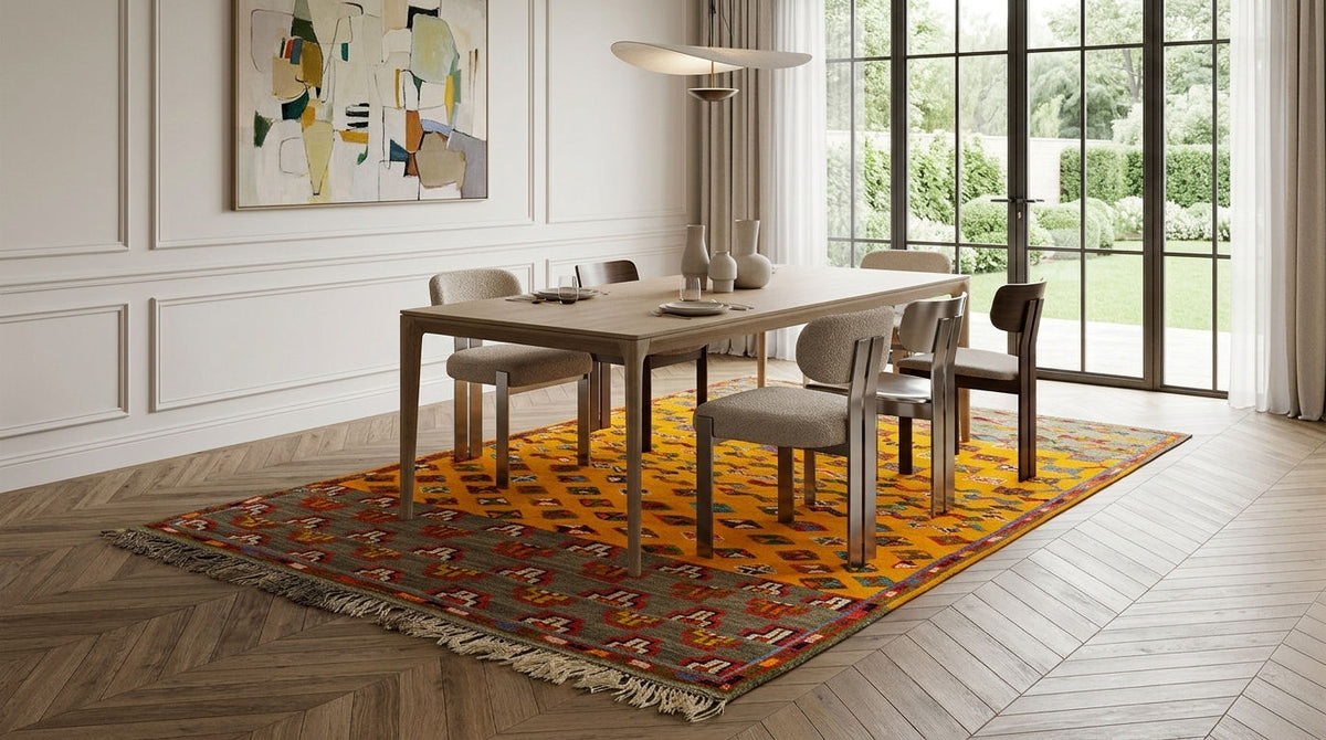 A dining room scene with a rectangular wooden dining table and six upholstered chairs placed on a large patterned area rug. The room has light wood herringbone flooring, white paneled walls, a wall art piece, a hanging pendant light, and tall windows looking out onto greenery.