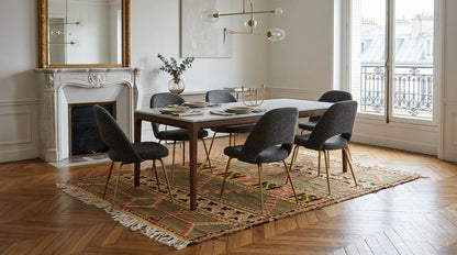 A dining room scene with a rectangular wooden table surrounded by six dark upholstered chairs on a patterned vintage moroccan kilim area rug, set in a bright room with a fireplace, large windows, and wood flooring.