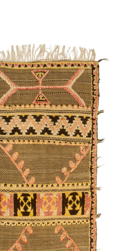 A close-up of a woven Moroccan  rug corner showing a geometric pattern in olive brown, black, pink, and beige with fringed edges on a white background.