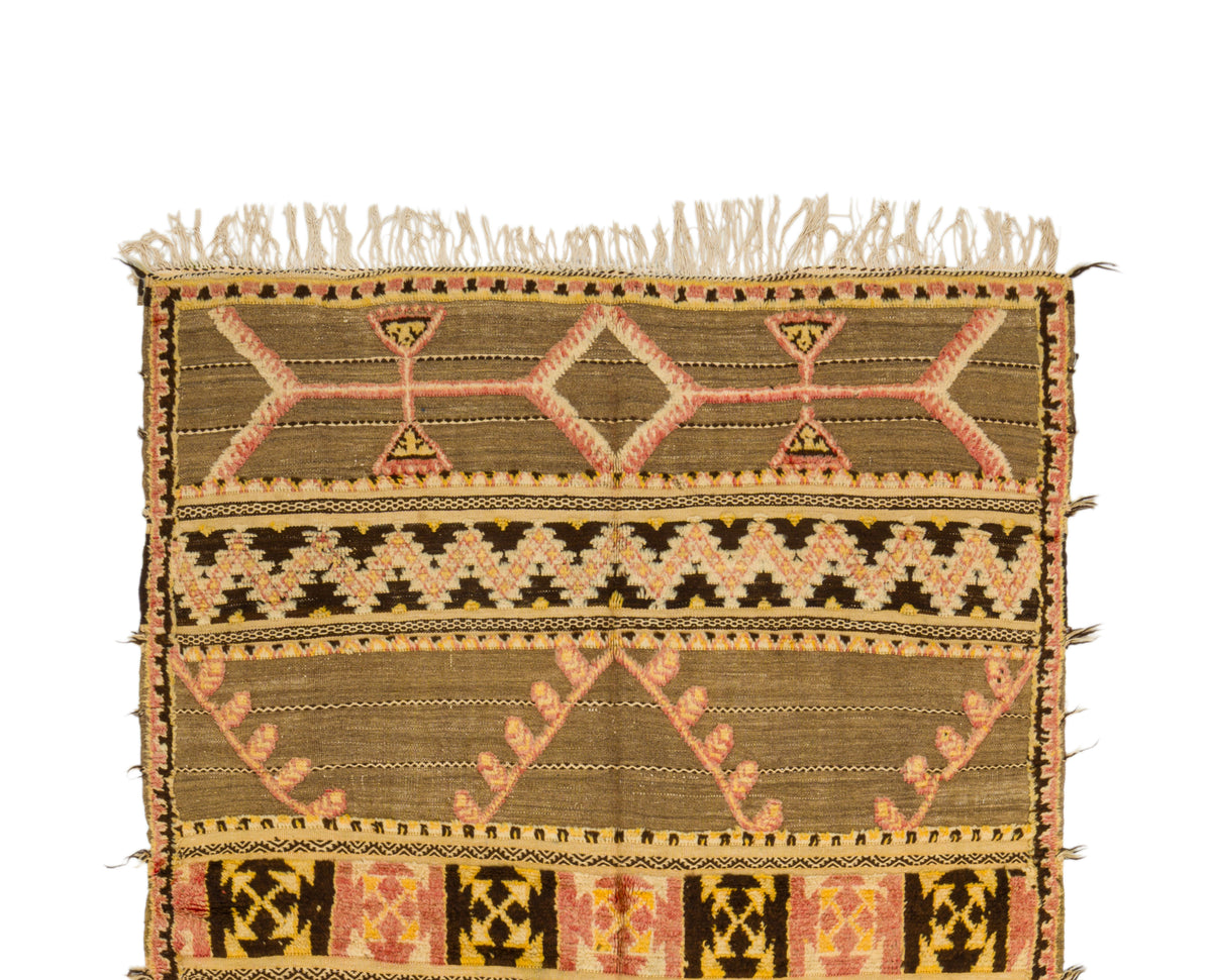A vintage moroccan kilim rug with fringe on the top edge, featuring geometric and tribal-inspired patterns in olive green, beige, pink, and black on a plain white background.