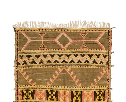 A vintage moroccan kilim rug with fringe on the top edge, featuring geometric and tribal-inspired patterns in olive green, beige, pink, and black on a plain white background.