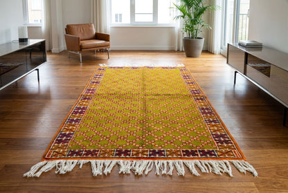 A rectangular Moroccan area rug with a yellow-green center, orange and brown patterned borders, and white fringe, displayed on a polished hardwood floor in a modern living room.