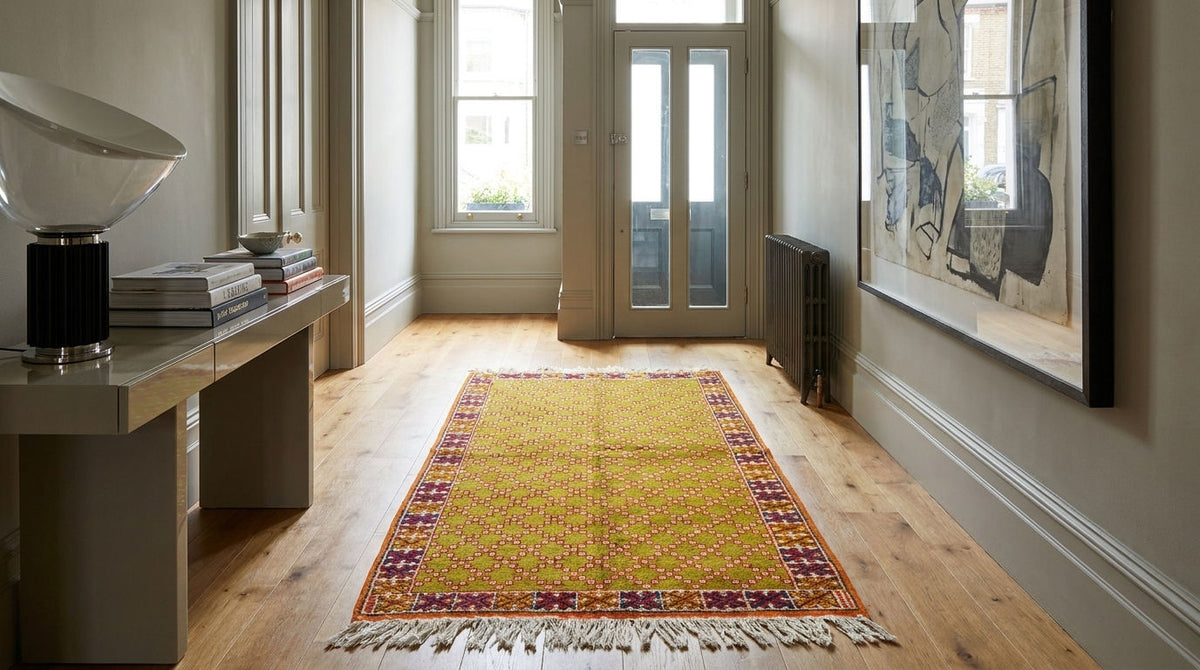 A rectangular yellow area rug with a red and blue patterned border and fringe, displayed on a light wood hallway floor.