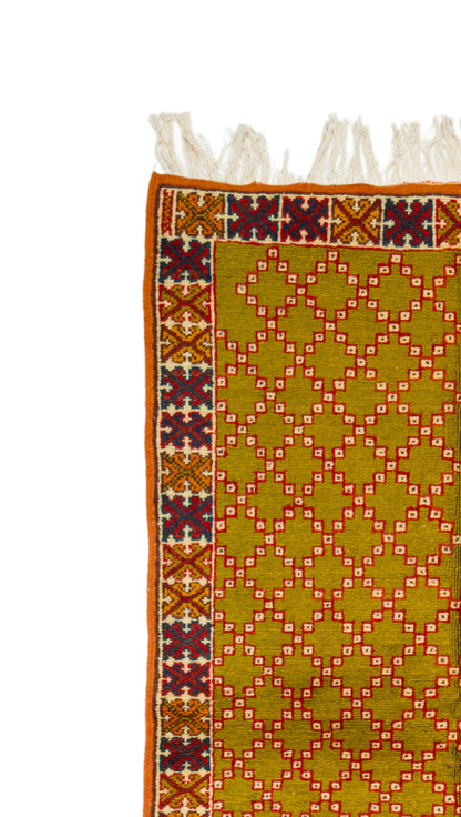 A close-up of a patterned rug showing a mustard yellow field with a repeating diamond lattice in red and white, bordered by a multicolor geometric pattern and white fringe tassels at the top.