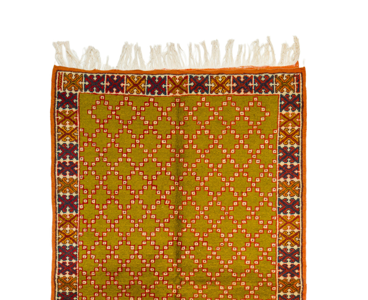 A Moroccan vintage rug with white fringe along the top edge, featuring a mustard yellow center with a repeating diamond lattice pattern in orange, red, and white, and a decorative border in orange, burgundy, and brown.