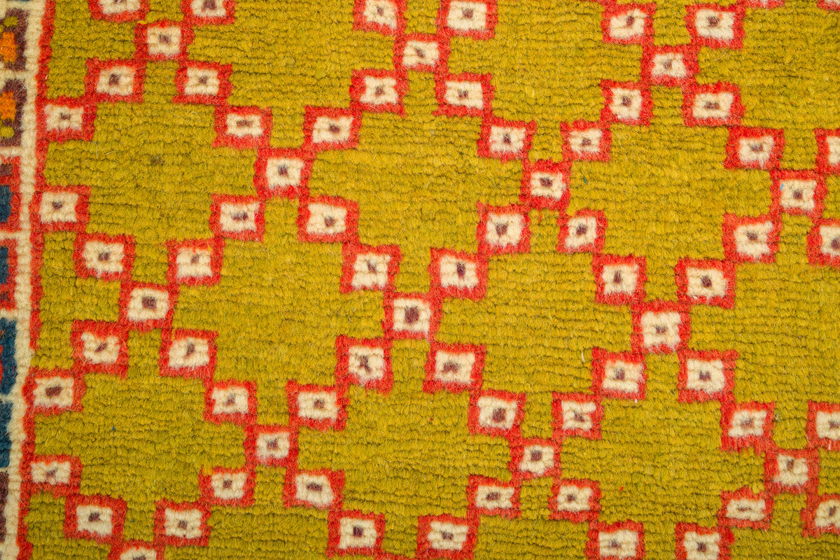 A close-up of a woven rug showing a mustard yellow base with a repeating red and cream diamond pattern.