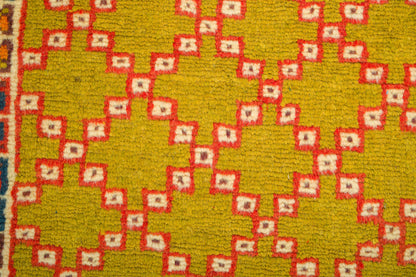 A close-up of a woven rug showing a mustard yellow base with a repeating red and cream diamond pattern.