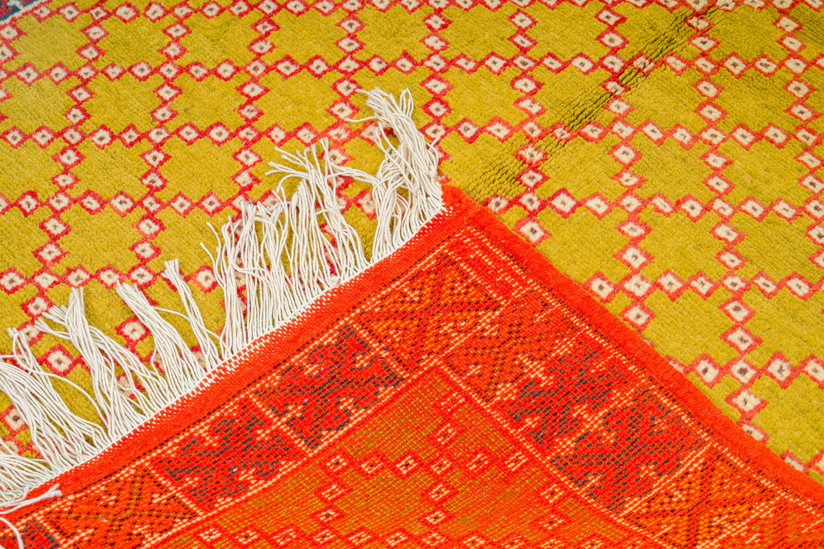 A close-up of a woven Moroccan rug with a yellow and red geometric pattern, white fringe, and a bright orange-red patterned corner.