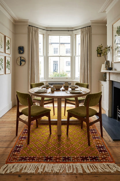 A round wooden dining table with four olive green upholstered chairs, set on a patterned orange and red area rug in a bright, traditional room with a bay window and fireplace.