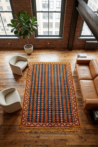 A rectangular area rug with a geometric pattern in navy, rust orange, and beige, styled on a wooden floor in a loft apartment with a leather sofa, armchairs, and a potted plant.