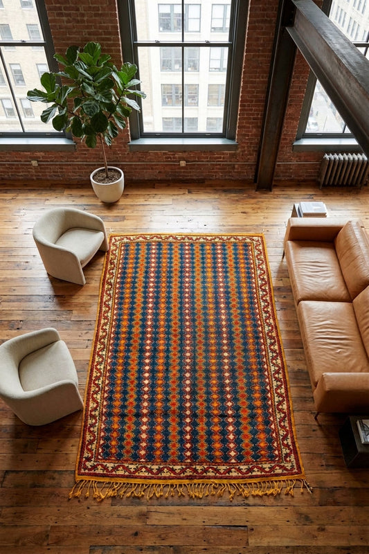 A rectangular area rug with a geometric pattern in navy, rust orange, and beige, styled on a wooden floor in a loft apartment with a leather sofa, armchairs, and a potted plant.