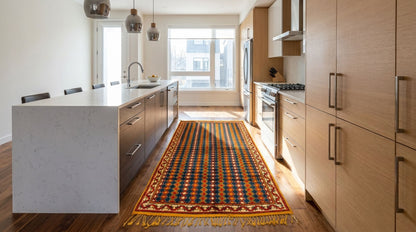 A long patterned area rug with fringe is displayed on a hardwood floor in a modern kitchen with light wood cabinets and a white island.