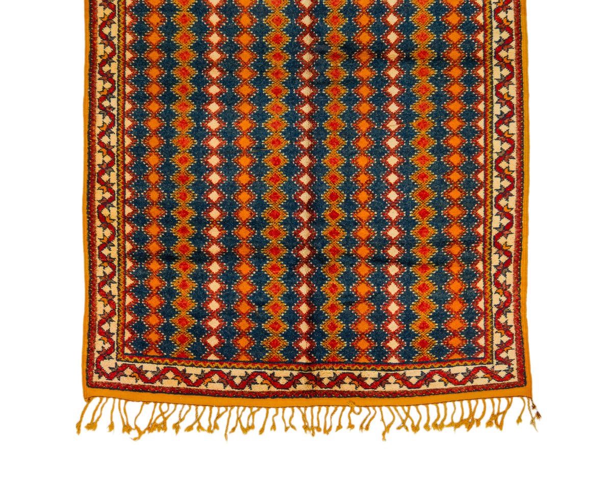 A square patterned rug with a geometric design in navy blue, orange, red, and cream, featuring a decorative border and fringe tassels along the bottom edge, shown against a plain white background.