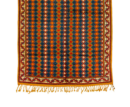 A square patterned rug with a geometric design in navy blue, orange, red, and cream, featuring a decorative border and fringe tassels along the bottom edge, shown against a plain white background.
