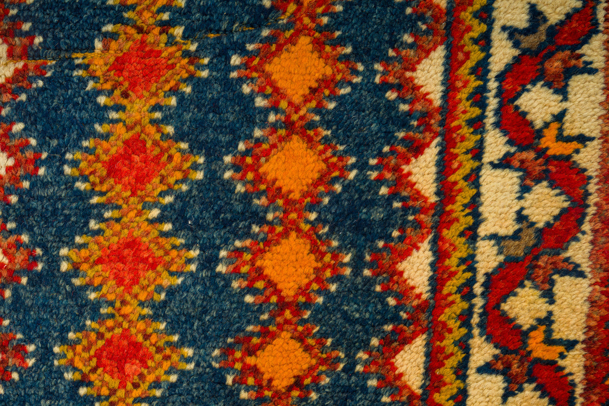 Close-up of a woven Moroccan rug showing a blue field with repeating orange and red diamond motifs and a cream border with red, orange, and blue patterning.