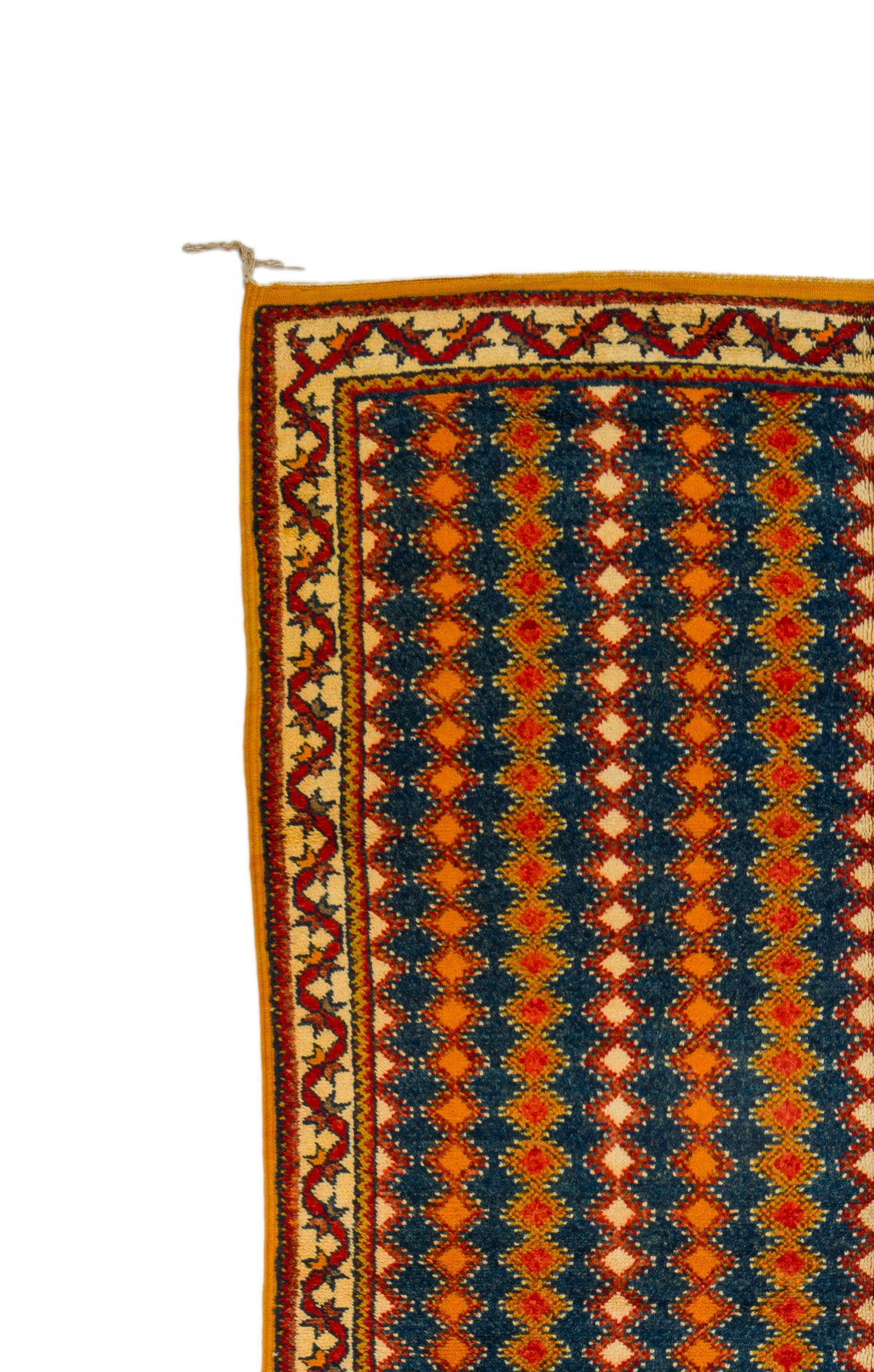 A close-up of a patterned rug showing a geometric design with navy blue, orange, red, and beige colors, featuring a decorative border and a small white hanging loop at the top left.
