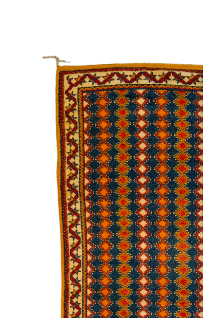 A close-up of a patterned rug showing a geometric design with navy blue, orange, red, and beige colors, featuring a decorative border and a small white hanging loop at the top left.