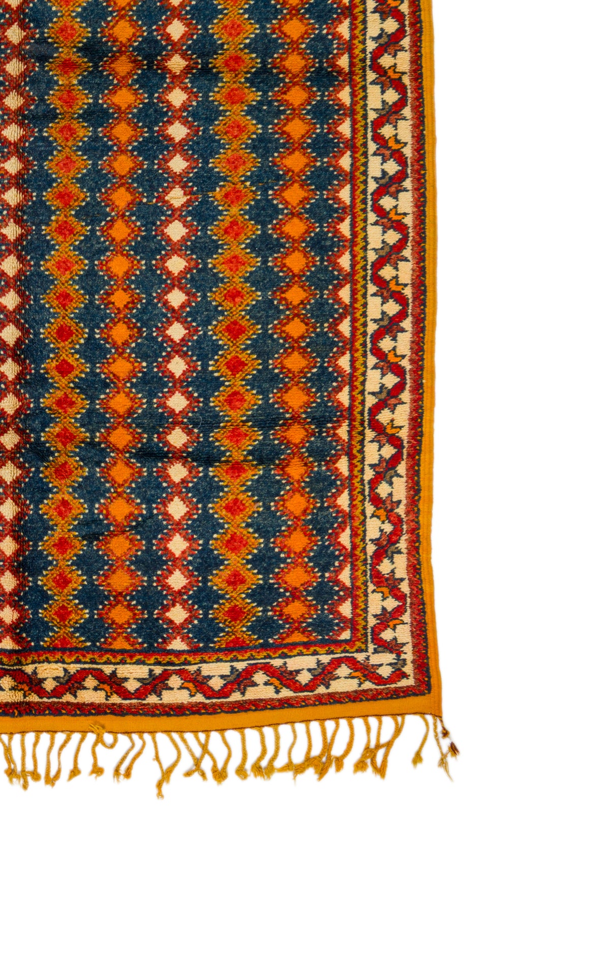 A close-up of a patterned rug corner showing a navy blue field with repeating orange and cream diamond motifs, a red and cream geometric border, and yellow fringe tassels on a white background.