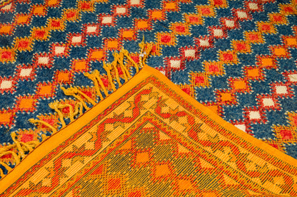 A close-up of a woven rug with a geometric pattern, showing a corner with yellow fringe. The rug features a blue field with repeating orange and white diamond motifs, bordered by a wide orange section with red and yellow geometric details.