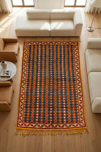 A rectangular area rug with a geometric diamond pattern in navy blue, rust orange, and cream, laid out on a light wood floor in a modern living room setting.