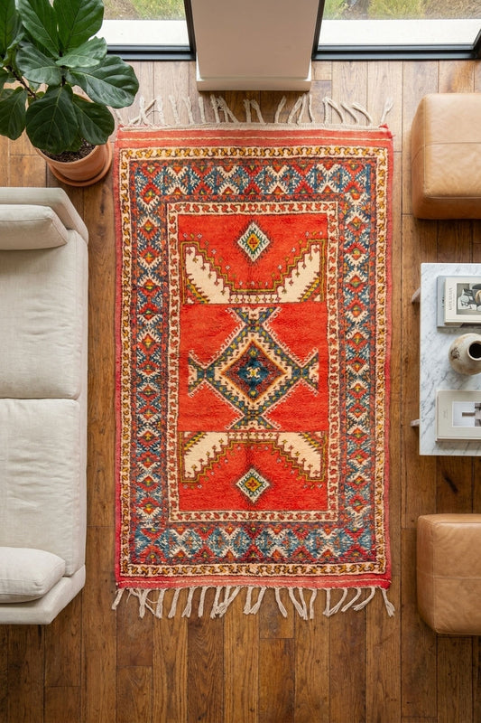 A rectangular red and orange patterned area rug from Morocco with cream, blue, and brown geometric motifs and fringe, shown hanging on a wooden floor in a modern living room setting.This one-of-a-kind 1970s vintage Moroccan wool runner rug was hand-knotted by a Berber woman artisan in the High Atlas Mountains of Morocco using traditional Taznakht rug-making techniques.