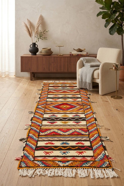 A long woven runner rug with a Southwestern geometric pattern in red, orange, yellow, blue, black, and cream, styled on a light wood floor in a modern living room setting.