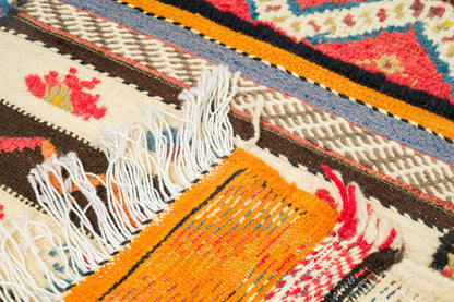 Close-up of a woven Moroccan rug with white fringe, featuring geometric and floral patterns in orange, cream, brown, red, blue, and black.