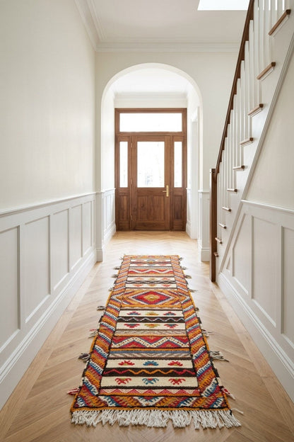 A long, colorful patterned Glaoui runner rug from Morocco with fringe is displayed on a light wood floor in a bright hallway with white wainscoting, a wooden staircase on the right, and a wooden front door at the end.