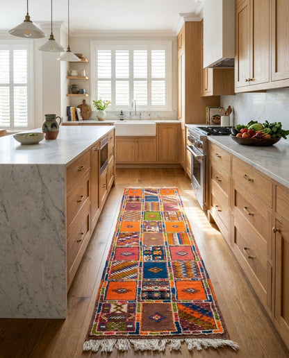 A colorful patchwork-style area rug with fringe is displayed on a hardwood floor in a bright, modern kitchen with light wood cabinets and marble countertops.