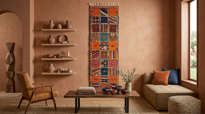A colorful patchwork kilim wall hanging with geometric patterns is displayed in a warm, bohemian-style living room with terracotta walls, a wooden armchair, and a sofa.