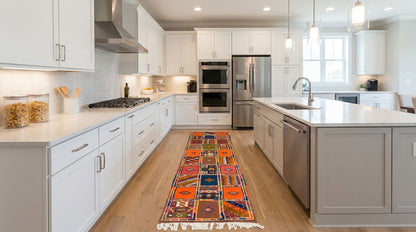 A long multicolor patterned runner rug with fringe is displayed on a light wood floor in a modern white kitchen with stainless steel appliances.