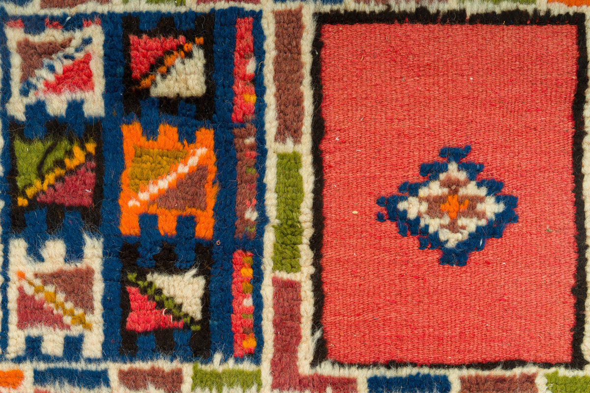 Close-up of a woven rug showing a coral red field with a central diamond motif in blue, white, and orange, alongside geometric panels in blue, orange, green, red, black, and cream.