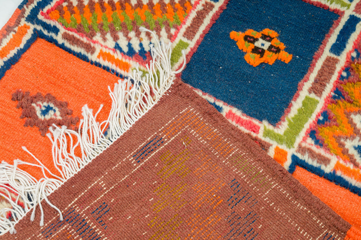 A close-up of a colorful woven rug with geometric patterns, showing orange, blue, brown, green, and cream sections with white fringe.