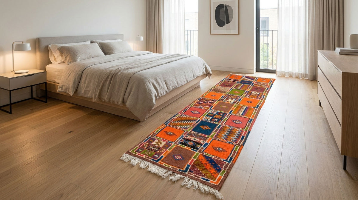 A colorful patchwork-style runner rug with white fringe is styled on a light wood floor in a modern, sunlit bedroom with a bed and a wooden dresser.