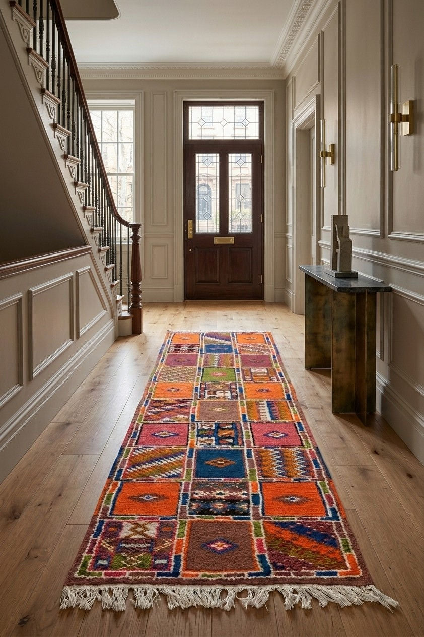 A long, multicolored patchwork-style runner rug with white fringe is displayed on a light wood hallway floor in an elegant interior setting with a staircase and console table.