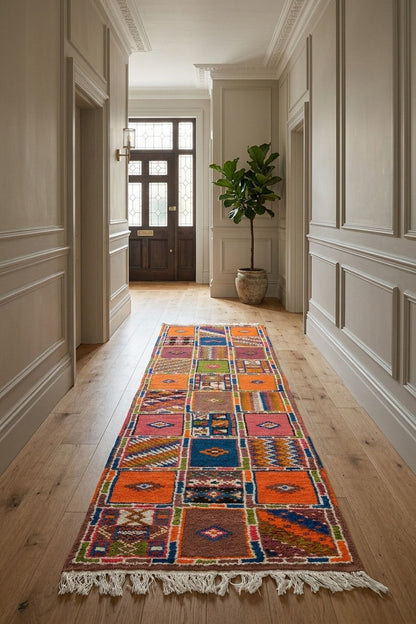 A long, narrow multicolor patchwork runner rug with fringe ends is displayed on a light wood hallway floor, leading towards a dark wooden door in the distance.
