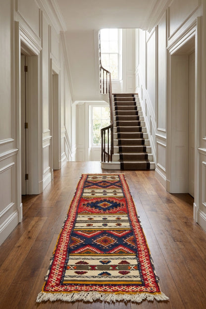 A long patterned wool runner rug with fringe, displayed on a hardwood floor in a bright hallway leading to a staircase.
