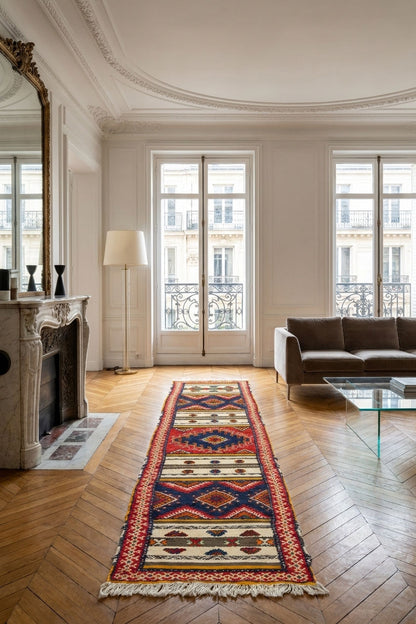A long rectangular patterned rug with fringe is displayed in a bright, elegant living room with parquet wood floors, a fireplace, and a sofa.