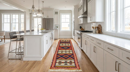 A long, colorful patterned runner rug is displayed on a light wood floor in a bright, modern white kitchen. The rug features a traditional geometric design with red, blue, yellow, and black patterns.