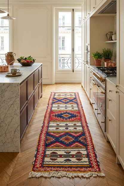 A long, narrow geometric patterned runner rug in red, blue, beige, and cream is displayed on a light wood herringbone floor in a bright, modern kitchen.