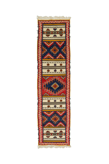 A long, narrow geometric runner rug with fringe on both ends, featuring a bold pattern of red, navy blue, beige, and black.