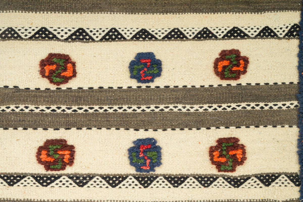 A close-up of a woven textile with horizontal bands, geometric diamond patterns, and stylized floral motifs in red, orange, green, blue, and brown on an off-white background.