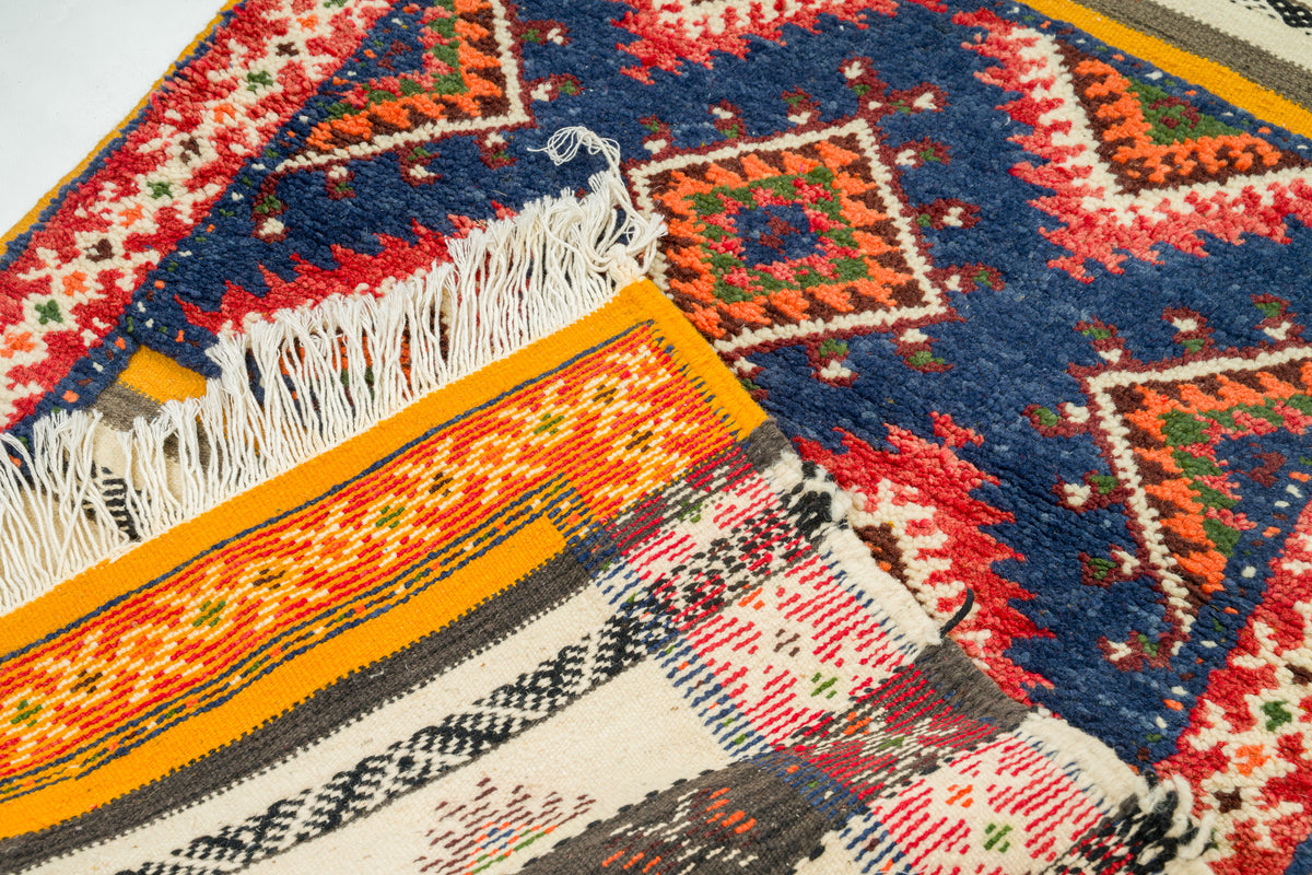 A close-up of a patterned woven rug with geometric motifs in navy blue, red, orange, cream, black, and white, including white fringe on one side.
