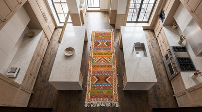 A colorful geometric patterned  Glaoui runner rug with fringe is displayed on a dark wood floor in a modern kitchen with white marble countertops and light wood cabinets.