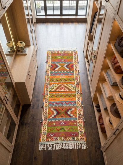 A long, narrow woven runner rug with a colorful geometric pattern and white fringe, displayed on a dark wood floor between tall light wood cabinets.